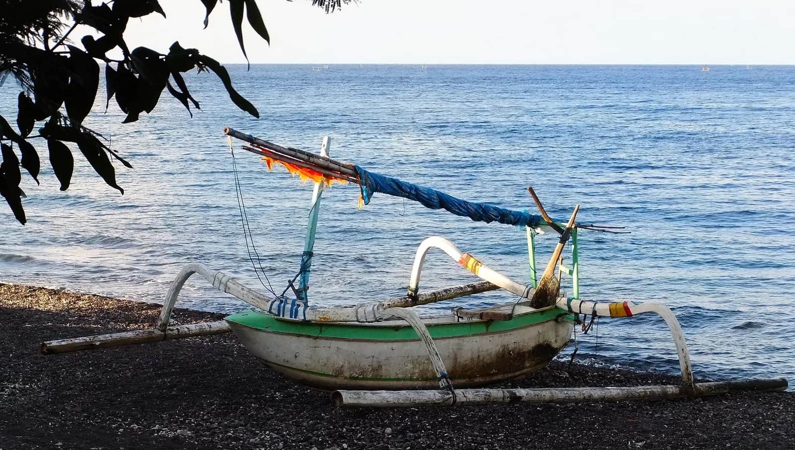 Jukung de pêcheur balinais à balancier échoué sur le rivage de galets noirs d'Amed face à une mer calme, est de Bali