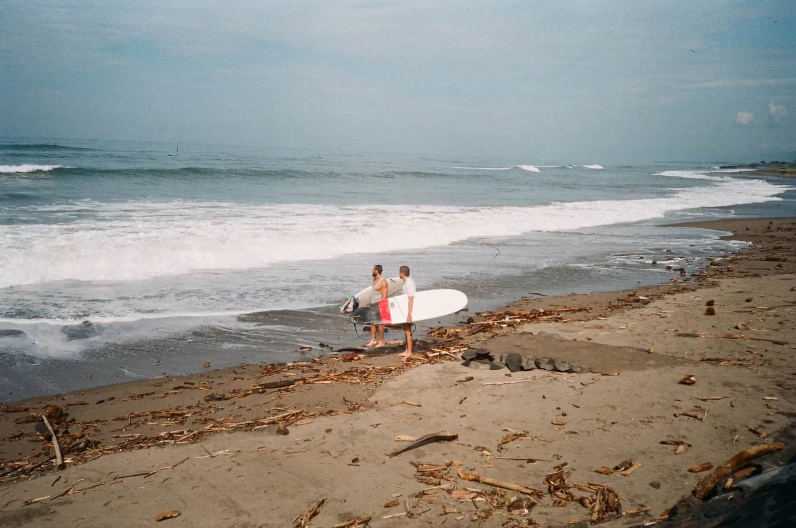 Deux surfeurs portant leurs longboards sur la plage de sable sombre de Batu Bolong à Canggu, Bali