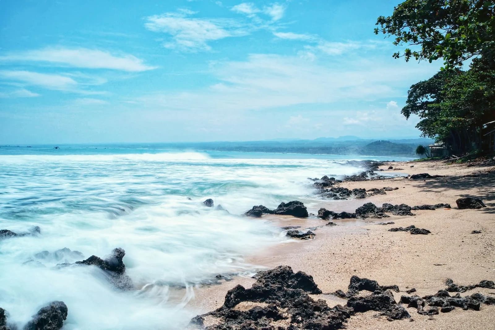 Plage de sable et de rochers volcaniques noirs bordée d'arbres, vagues déferlantes, côte de Java occidental