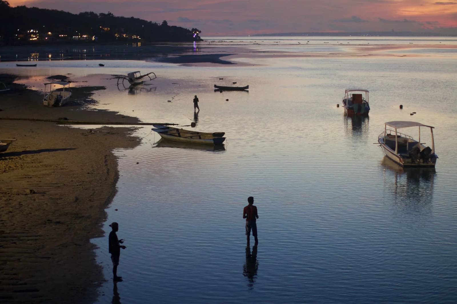 Marée basse au petit matin sur une plage : pêcheurs en silhouette dans l'eau peu profonde et bateaux ancrés